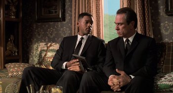 Movie still from “Men in Black” (1997), directed by Barry Sonnenfeld – Two men in suits sitting next to each other on a couch; Medium shot, Over the shoulder angle