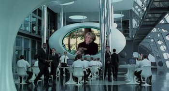 Movie still from “Men in Black II” (2002), directed by Barry Sonnenfeld – A group of people are gathered around a round table; Wide shot, Over the shoulder angle