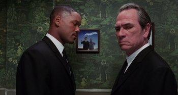 Movie still from “Men in Black II” (2002), directed by Barry Sonnenfeld – Two men in suits standing next to each other in front of a picture; Medium shot, Over the shoulder angle