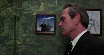 Movie still from “Men in Black II” (2002), directed by Barry Sonnenfeld – A man in a suit and tie next to a picture of two men; Medium shot, Over the shoulder angle