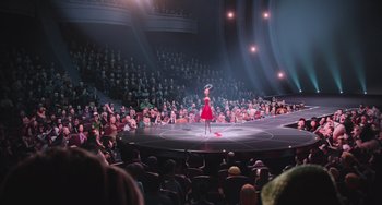 Movie still from “Minions” (2015), directed by Pierre Coffin – A woman in a red dress is on a stage in front of an audience; Extreme Wide shot, High angle