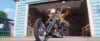 Movie still from “Minions: The Rise of Gru” (2022), directed by Jonathan del Val – A man sitting on the back of a motorcycle in front of a garage door; Wide shot, Low angle