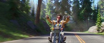 Movie still from “Minions: The Rise of Gru” (2022), directed by Jonathan del Val – A man riding a motorcycle down a road; Wide shot, Low angle