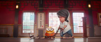 Movie still from “Minions: The Rise of Gru” (2022), directed by Jonathan del Val – A woman is holding a pair of scissors next to a minion in front of a window; Wide shot, Low angle