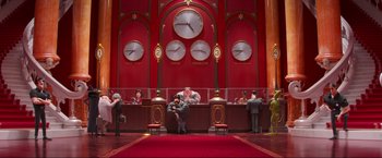 Movie still from “Minions: The Rise of Gru” (2022), directed by Jonathan del Val – A group of people sitting in a room with a clock on the wall behind them; Wide shot, Low angle