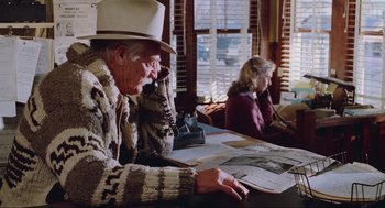 Movie still from “Misery” (1990), directed by Rob Reiner – An older man sitting at a table while a woman sits at a desk; Medium shot, High angle