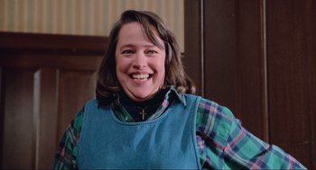 Movie still from “Misery” (1990), directed by Rob Reiner – A woman smiling for the camera wearing an apron and plaid shirt; Medium shot, Low angle