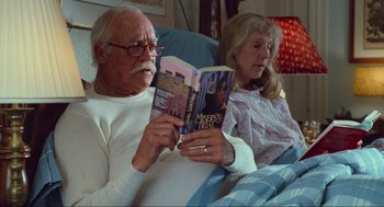 Movie still from “Misery” (1990), directed by Rob Reiner – An older man and woman sitting in a chair reading a magazine; Medium shot, High angle