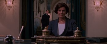 Movie still from “Mission: Impossible” (1996), directed by Brian De Palma – An older woman sitting at a desk in front of a man in a suit; Medium shot, Low angle
