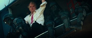 Movie still from “Mission: Impossible III” (2006), directed by J.J. Abrams – A man in a suit and tie sitting next to a statue; Medium shot, Low angle