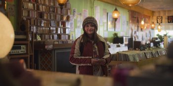 Movie still from “Mixtape” (2021), directed by Valerie Weiss – A woman standing in front of a table in a room; Medium shot, Over the shoulder angle