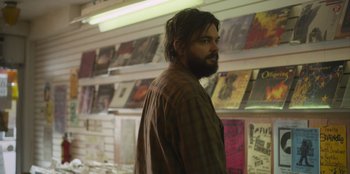 Movie still from “Mixtape” (2021), directed by Valerie Weiss – A man with a beard standing next to a wall of records; Medium shot, Over the shoulder angle