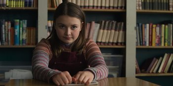 Movie still from “Mixtape” (2021), directed by Valerie Weiss – A girl sitting at a table in front of a book case; Medium shot, Low angle