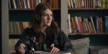 Movie still from “Mixtape” (2021), directed by Valerie Weiss – A young woman sitting at a table in front of a book shelf; Close Up shot, Over the shoulder angle