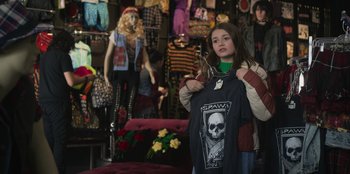 Movie still from “Mixtape” (2021), directed by Valerie Weiss – A young girl holding up a t - shirt in front of mannequins; Medium shot, Over the shoulder angle
