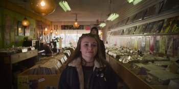 Movie still from “Mixtape” (2021), directed by Valerie Weiss – A woman standing in front of a bunch of tables; Medium shot, Over the shoulder angle