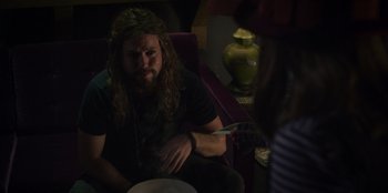 Movie still from “Mixtape” (2021), directed by Valerie Weiss – A man sitting on a chair with a beard and long curly hair; Medium shot, Over the shoulder angle