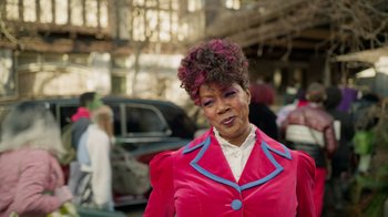 Movie still from “Monster High 2” (2023), directed by Todd Holland – An older woman in a pink jacket is standing in front of a crowd; Medium shot, Over the shoulder angle