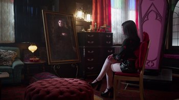 Movie still from “Monster High 2” (2023), directed by Todd Holland – A woman sitting in front of a painting in a room; Wide shot, Over the shoulder angle