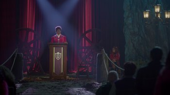 Movie still from “Monster High 2” (2023), directed by Todd Holland – A man in a red jacket is speaking at a podium in front of people; Wide shot, Over the shoulder angle