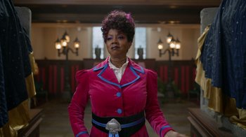 Movie still from “Monster High 2” (2023), directed by Todd Holland – A woman in a pink coat standing in front of a wall; Medium shot, Low angle