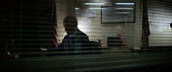 Movie still from “Monsters of Man” (2020), directed by Mark Toia – A man sitting at a desk in front of an american flag; Medium shot, Low angle