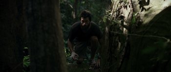 Movie still from “Monsters of Man” (2020), directed by Mark Toia – A man crouching down in the woods near a tree; Wide shot, Low angle