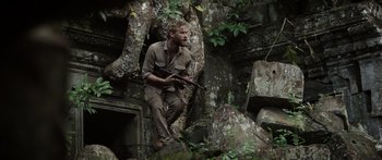Movie still from “Monsters of Man” (2020), directed by Mark Toia – A man sitting on top of a rock holding a rifle; Medium shot, Low angle