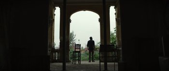 Movie still from “Monsters of Man” (2020), directed by Mark Toia – A man standing in front of two chairs in an archway; Wide shot, Low angle