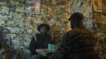 Movie still from “Atlanta” (2016), created by Donald Glover – Two men are sitting at a table in front of a wall covered in stickers; Medium shot, Over the shoulder angle