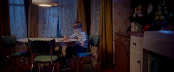 Movie still from “Monstrous” (2022), directed by Chris Sivertson – A young boy sitting at a table in a room; Wide shot, High angle