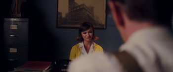 Movie still from “Monstrous” (2022), directed by Chris Sivertson – A woman in a white shirt and a yellow jacket is in front of a painting; Medium shot, Over the shoulder angle