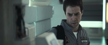 Movie still from “Moon” (2009), directed by Duncan Jones – A man in a gray and orange suit is looking at boxes; Close Up shot, Over the shoulder angle