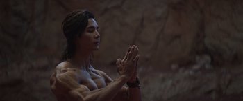 Movie still from “Mortal Kombat” (2021), directed by Simon McQuoid – A man with long black hair is praying in a cave; Medium shot, Low angle