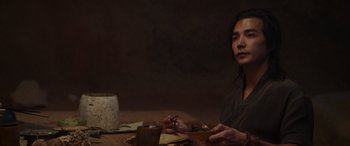Movie still from “Mortal Kombat” (2021), directed by Simon McQuoid – A person sitting at a table with a cup of coffee; Medium shot, Over the shoulder angle