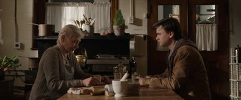 Movie still from “Mr. Harrigan's Phone” (2022), directed by John Lee Hancock – Two people sitting at a table in front of a fireplace; Medium shot, Over the shoulder angle