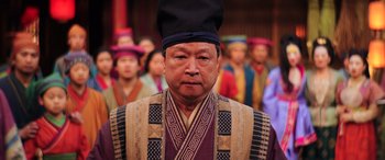 Movie still from “Mulan” (2020), directed by Niki Caro – An older man wearing a black hat in front of a group of people; Close Up shot, Low angle