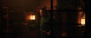 Movie still from “Mulan” (2020), directed by Niki Caro – A man holding a sword while standing next to a lamp; Medium shot, Over the shoulder angle