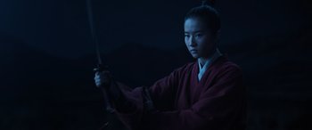 Movie still from “Mulan” (2020), directed by Niki Caro – A woman holding a sword in front of a dark background; Medium shot, Low angle