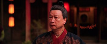 Movie still from “Mulan” (2020), directed by Niki Caro – An older asian man wearing a red and black robe; Close Up shot, Low angle