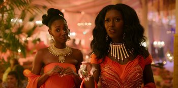 Movie still from “Murder Mystery 2” (2023), directed by Jeremy Garelick – Two beautiful women in orange dresses and gold jewelry; Close Up shot, Over the shoulder angle