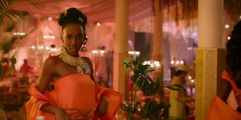 Movie still from “Murder Mystery 2” (2023), directed by Jeremy Garelick – A woman in an orange dress standing next to a plant; Medium shot, Over the shoulder angle