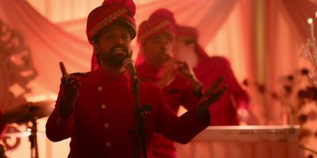 Movie still from “Murder Mystery 2” (2023), directed by Jeremy Garelick – A man wearing a red turban is speaking into a microphone; Medium shot, Over the shoulder angle