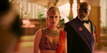 Movie still from “Murder Mystery 2” (2023), directed by Jeremy Garelick – A woman in a red and orange outfit and a man in a suit; Medium shot, Over the shoulder angle