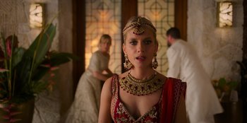 Movie still from “Murder Mystery 2” (2023), directed by Jeremy Garelick – A woman in a red and gold dress and a red and gold headpiece; Medium shot, Over the shoulder angle