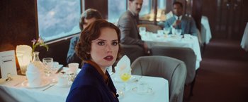 Movie still from “Murder on the Orient Express” (2017), directed by Kenneth Branagh – A woman sitting at a table in front of a glass of orange juice; Medium shot, Over the shoulder angle