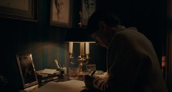 Movie still from “My Policeman” (2022), directed by Michael Grandage – A man writing on a piece of paper in a dimly - lit room; Medium shot, High angle
