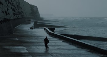 Movie still from “My Policeman” (2022), directed by Michael Grandage – A man walking a dog down a sidewalk next to the ocean; Extreme Wide shot, Low angle