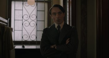 Movie still from “My Policeman” (2022), directed by Michael Grandage – A man wearing a suit and tie standing with his arms crossed; Medium shot, Over the shoulder angle