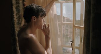 Movie still from “My Policeman” (2022), directed by Michael Grandage – A man sitting in front of a window looking out at a river; Medium shot, High angle